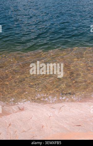 L'acqua cristallina del lago si snoda su un bordo di sabbia poco profondo accanto alla roccia rosa liscia, creando una tranquilla scena costiera naturale in piena luce del giorno. Foto Stock