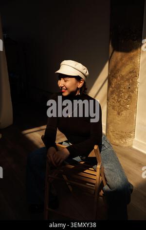 Una donna è seduta su una sedia di legno. Indossa un cappello e si siede in un'area illuminata dal sole di una stanza. La scena mostra una luce soffusa e uno sfondo semplice con un co Foto Stock
