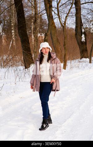 Una donna cammina lungo un sentiero innevato in una foresta indossa un cappotto caldo e alberi di cappello la circondano mentre si gode il filtro della luce solare fredda del giorno invernale Foto Stock