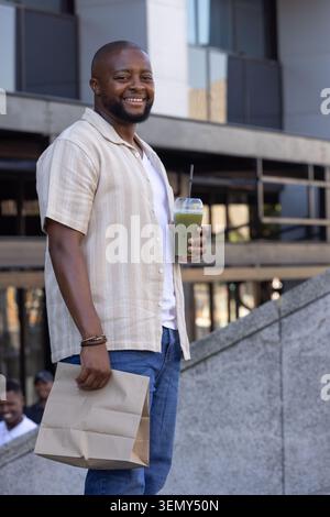 Uomo africano adulto sorridente, con bevanda verde, paglia nera, sacchetto di carta su gradini di pietra fuori dalla piazza Foto Stock