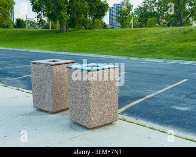 Due bidoni della spazzatura si trovano in un parcheggio accanto a un'area erbosa con alberi. Sono vuote e collocate sul marciapiede. Edifici alti possono essere visti nel di Foto Stock