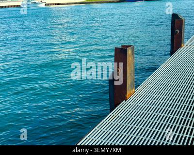 Una banchina in metallo si estende sull'acqua limpida. Le barche sono viste nelle vicinanze. Il sole splende, riflettendo sulla superficie. La terra è presente nel backgroun Foto Stock