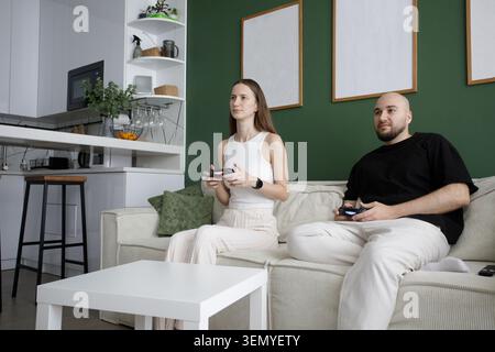 Allegra giovane coppia felice che gioca ai videogiochi sul divano in un moderno interno bianco e verde a casa Foto Stock