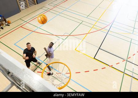 Diversi giocatori maschili in maglie che saltano e raggiungono il pallacanestro volando vicino al cerchio in palestra Foto Stock