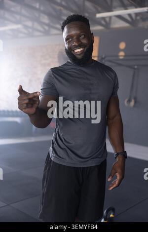 Uomo afro-americano in palestra sorridente, punta, indossa camicia aderente, campanello e anelli Foto Stock