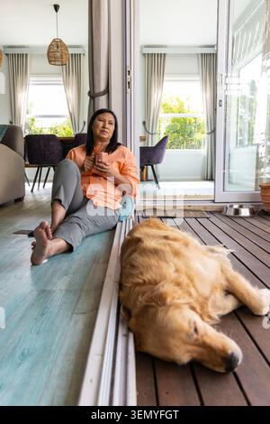 Donna asiatica di mezza età seduta davanti a una porta scorrevole di vetro che tiene la tazza di ceramica, cane che dorme fuori Foto Stock