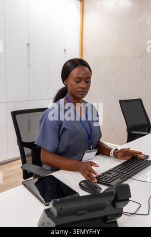 Donna africana in scrub grigi che indossa un cordone blu che digita la tastiera alla reception della clinica Foto Stock