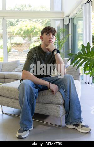 Adolescente seduto su un ottomano grigio in soggiorno, pensando, guardando vicino a Monstera e guardando Foto Stock