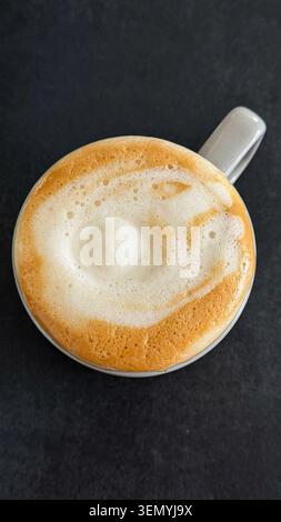 Vista dall'alto di una tazza di caffè in ceramica bianca riempita di cappuccino schiumoso su una superficie scura Foto Stock