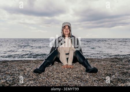 Donna da spiaggia ushanka giovane donna che indossa una giacca di pelle e un cappello di pelliccia si siede su una spiaggia di ciottoli in riva al mare sotto il cielo nuvoloso Foto Stock