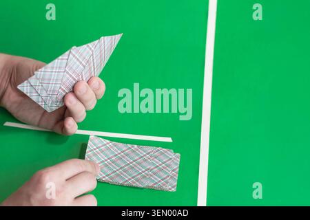 Le mani distribuiscono le carte da gioco con i dorsi a quadri su un tavolo verde con linee bianche. La mano sinistra tiene la ventola delle carte, la mano destra prende la carta dal mazzo. Dita e pa Foto Stock