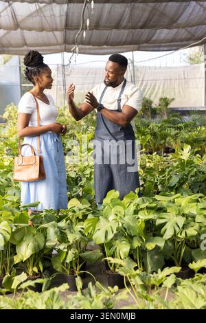 Uomo e donna afro-americana, uomo in grembiule grigio gestante, donna che tiene un sacchetto abbronzante tra le piante Foto Stock