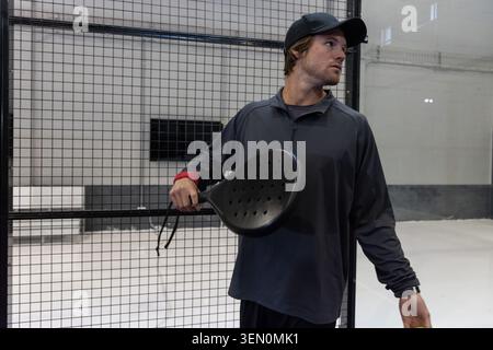 Uomo adulto in piedi all'interno del campo di padel vicino a una recinzione in rete metallica che indossa un berretto con racchetta perforata Foto Stock