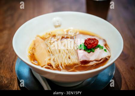 Ciotola di ramen con spaghetti, carne a fette, cipolle verdi e un pomodoro su un tavolo di legno. Foto Stock