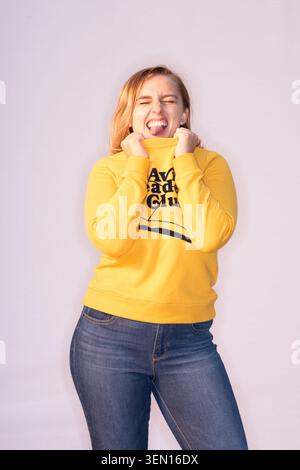 Hannah Witton si trova in uno studio con indosso una felpa gialla con un sorriso. Sta facendo una faccia giocosa mentre tiene la felpa. Foto Stock