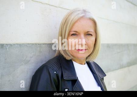 Donna con capelli biondi corti che indossa una giacca nera su uno sfondo in pietra chiara. Foto Stock