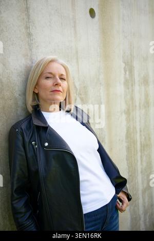 Donna bionda in una giacca in pelle si appoggia a una parete di cemento per un'espressione sicura. Foto Stock