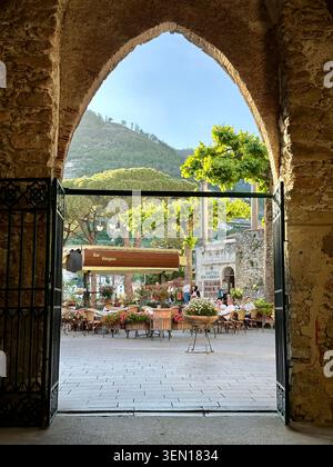 Un arco di pietra appuntito si apre su una piazza del villaggio illuminata dal sole, incorniciando tavoli da caffè, fiori e colline verdi oltre Foto Stock
