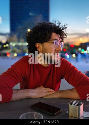 Giovane uomo in maglione rosso con occhiali seduto al tavolo all'aperto nel ritratto serale della città Foto Stock