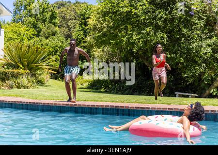 Amici afro-americani che spruzzano, ridono nella piscina del cortile sul galleggiante di ciambella rosa con delle sfumature Foto Stock