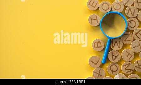 Una lente d'ingrandimento blu appoggiata su una pila di lettere alfabetiche di legno rotonde disposte sul lato destro di una superficie gialla pulita. Foto Stock