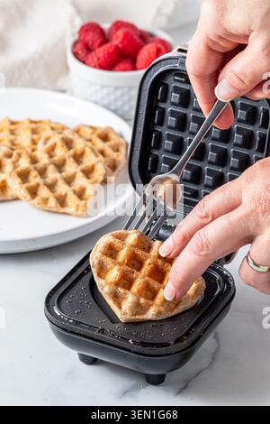 Un primo piano di una mano che rimuove i waffle appena fatti da una mini macchina per waffle. Foto Stock