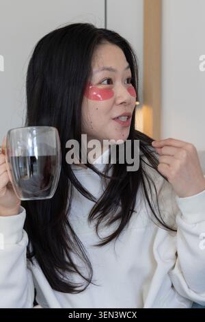 Donna asiatica che indossa una felpa che tiene la tazza a doppia parete con toppe rosse agli occhi, toccando i capelli a casa Foto Stock