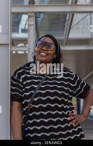 Donna afroamericana appoggiata alla colonna e sorridente sotto la tettoia di vetro con occhiali da sole Foto Stock