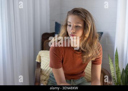 Seduto in una poltrona in rattan nel soggiorno, donna adulta che indossa un top a coste ruggine e pantaloni verdi Foto Stock
