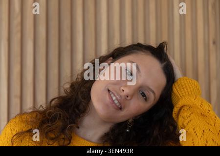 Donna adulta che inclina la testa, sorride contro le spranghe di legno, indossa un maglione giallo senape, orecchini a cerchio Foto Stock