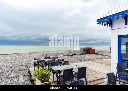 Tavoli e sedie scuri disposti sul ponte di legno di un caffè sul mare sulla spiaggia di ciottoli. A destra è visibile una struttura bianca con dettagli blu Foto Stock