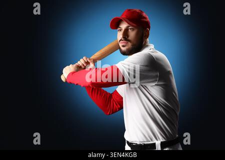 Determinato giocatore di baseball con cappello rosso e maglia bianca che tiene la mazza pronta a dondolare su sfondo blu scuro, l'atleta mostra concentrazione e concentrazione Foto Stock