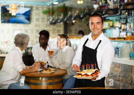 Sorridente cameriere latino nel grembiule che serve antipasti, tapas, spuntini sugli spiedini per persone diverse nel pub Foto Stock