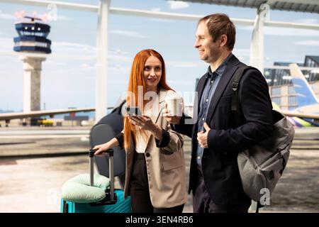 Uomo e donna sorridenti che chiacchierano in aeroporto, tenendo in mano una tazza di caffè, uno smartphone e un bagaglio. Viaggiatori d'affari felici che trasportano bagagli nel moderno terminal dell'aeroporto, parlano e utilizzano il telefono cellulare Foto Stock