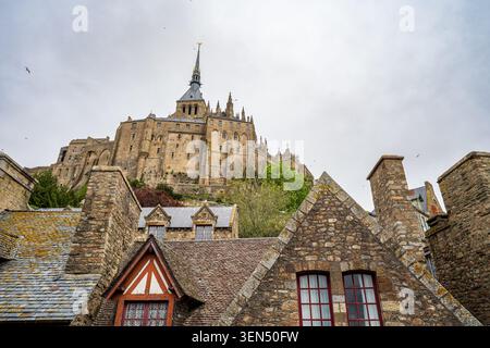 Una vista dall'angolo basso mette in risalto i tetti in ardesia e gli alti camini di pietra del villaggio di Mont Saint Michel in Normandia. Sullo sfondo, l'imponente G. Foto Stock
