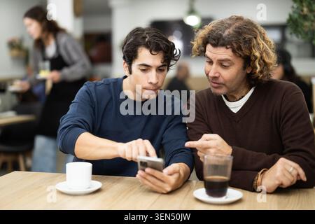 Due amici maschi al bar sono seduti al tavolo e guardano la foto sul loro telefono Foto Stock