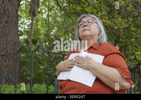 Donna anziana che si gode momenti tranquilli nella natura, tiene un libro, splende il sole sul viso. Contemplazione e tranquillità Foto Stock