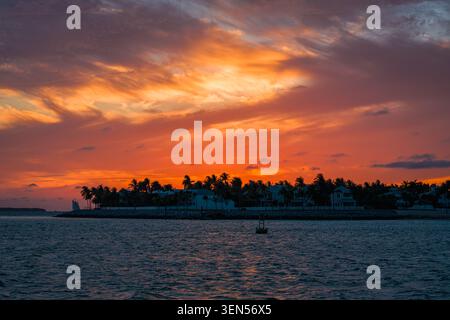 La piccola barca a vela per passeggeri attraversa l'oceano scuro vicino alla sagoma dell'isola di Key West sotto le formazioni di nuvole rosse durante il crepuscolo. Foto Stock