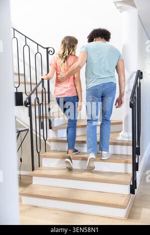 Indossate un abbigliamento casual che cammina su una scala di legno a casa, un uomo che mette il braccio intorno alla donna Foto Stock