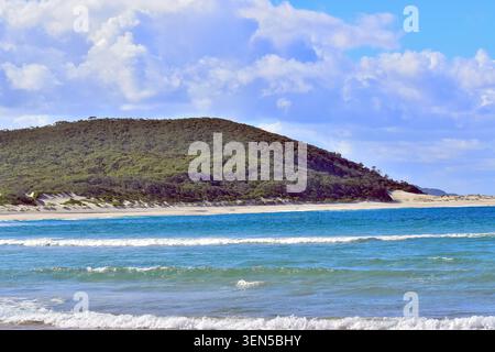 Le dolci onde turchesi si infrangono su una spiaggia di sabbia leggera, incontrando una lussureggiante collina boscosa che si innalza ripidamente dietro la costa. Foto Stock