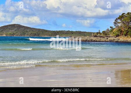 Le onde dolci si infrangono su una spiaggia di sabbia pallida, che conduce l'occhio verso un promontorio roccioso coperto da una lussureggiante vegetazione verde. Dietro di essa, sorgono colline boscose Foto Stock