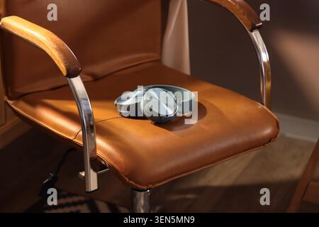 Le moderne cuffie wireless grigie su sedia da ufficio in pelle marrone, la tecnologia audio e di ascolto musicale, la comoda poltrona e le cuffie eleganti lo consigliano Foto Stock