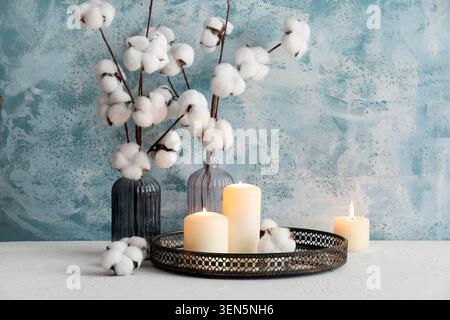 Accogliente arredamento casalingo con rami di cotone in vasi e candele accese su un vassoio decorativo contro una parete blu, la disposizione si trova su una superficie bianca agains Foto Stock