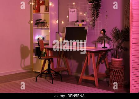 Moderno spazio di lavoro per la casa con monitor per computer, scaffale e luce arcobaleno al neon con illuminazione ambiente rosa Foto Stock