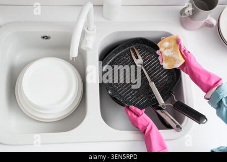 Persona con guanti in gomma rosa che lava la padella sporca con spugna e sapone nel lavello della cucina, nelle faccende domestiche e nella routine di pulizia Foto Stock