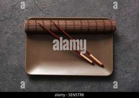 Piatto rettangolare marrone vuoto con bacchette di legno scuro e tappetino di bambù su sfondo grigio testurizzato, questo set è pronto per un pasto asiatico, come previsto Foto Stock