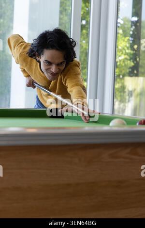 Uomo indiano in maglione di senape appoggiato sul tavolo da biliardo a casa, puntando sulle palle bianche e rosse Foto Stock