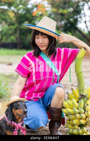 Cappello da donna banana cane giardino tradizionale abbigliamento rosa ritratto all'aperto donna sorridente che indossa un cappello di paglia e un abbigliamento tradizionale rosa siede nel giardino Be Foto Stock