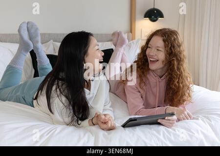 Diverse amiche distese su un letto bianco, di fronte all'altro e ridendo, tenendo un tablet Foto Stock