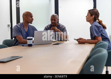 Team medico diversificato che indossa scrub discutere caso in sala conferenze con laptop, smartphone. Collaborazione, riunione, clinica, tablet, notebook, tazza Foto Stock